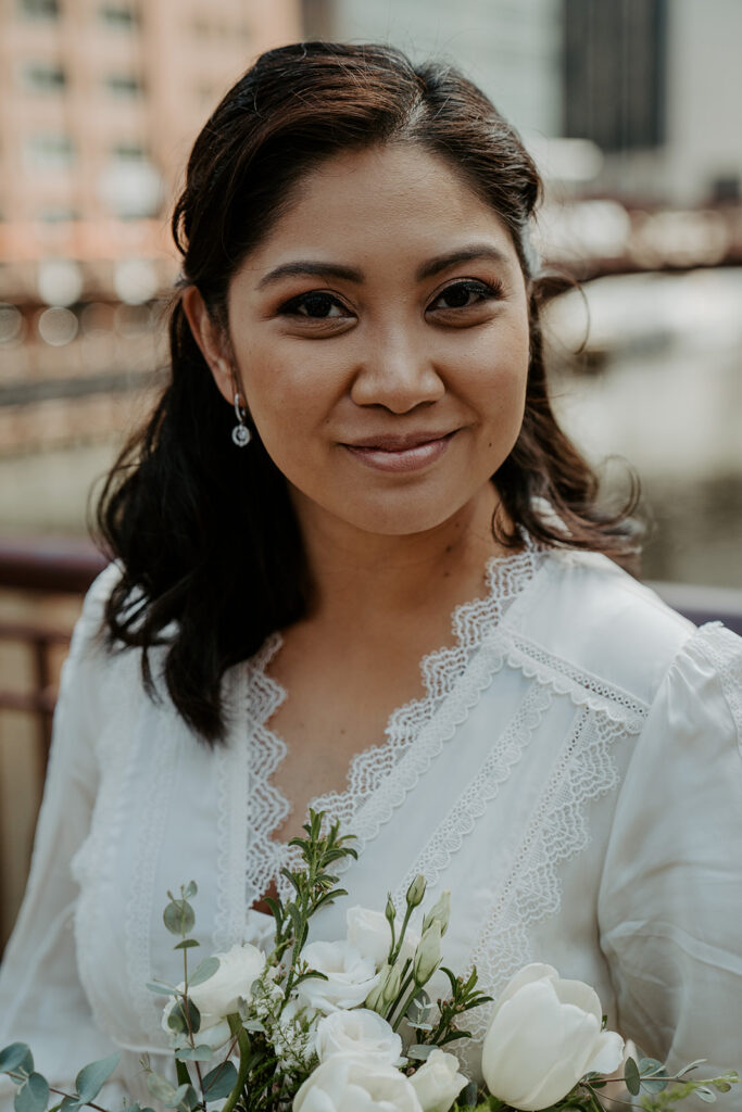 A bride smiles at the camera on her wedding day.  She wears a lacy white dress and silver earrings