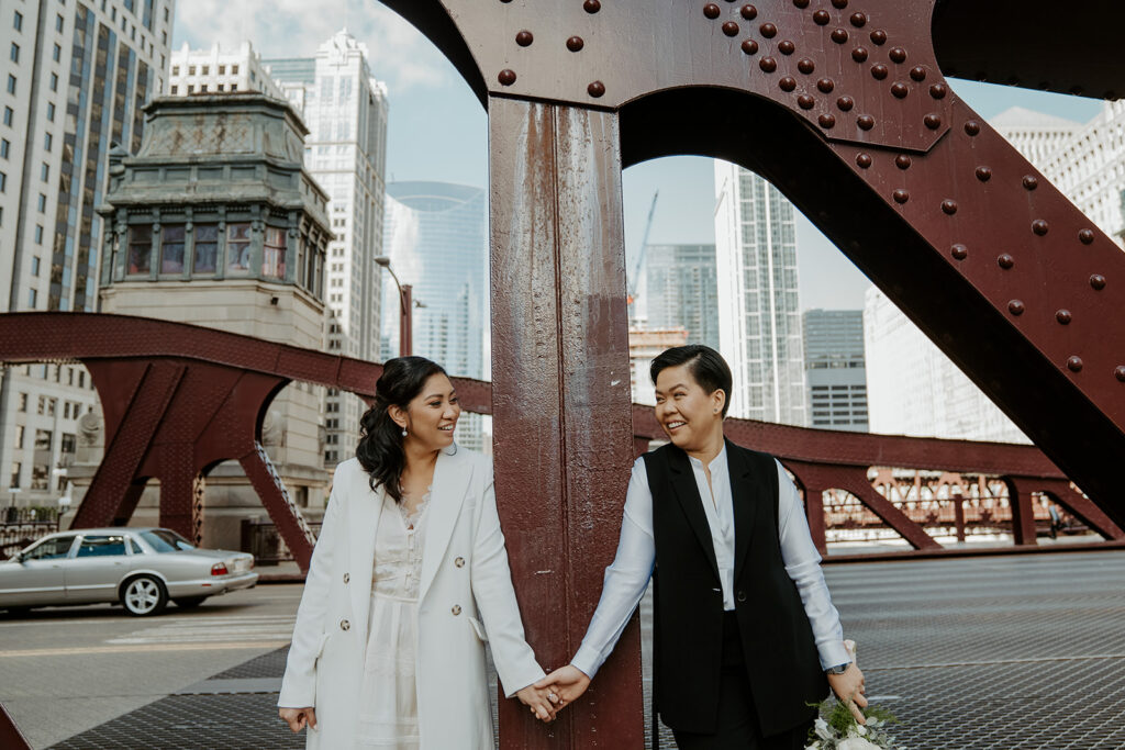 Two brides stand on Dusable bridge 
 leaning against one of its iconic pillars for their wedding portraits in the city of Chicago after their City Hall elopement.