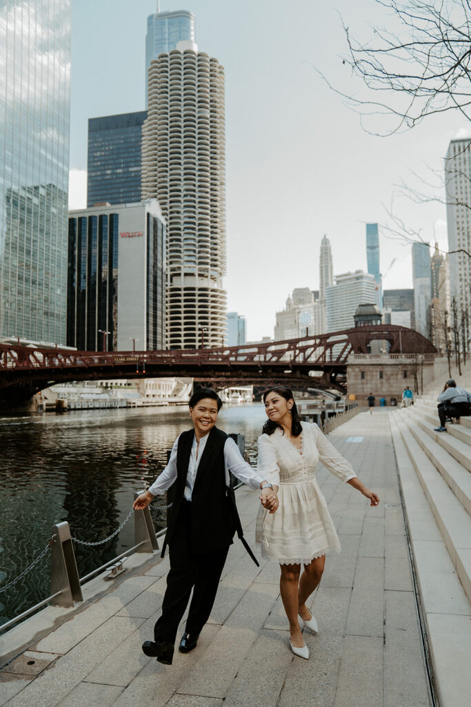Two brides walk together on the River Walk in Chicago holding hands and smiling at one another.