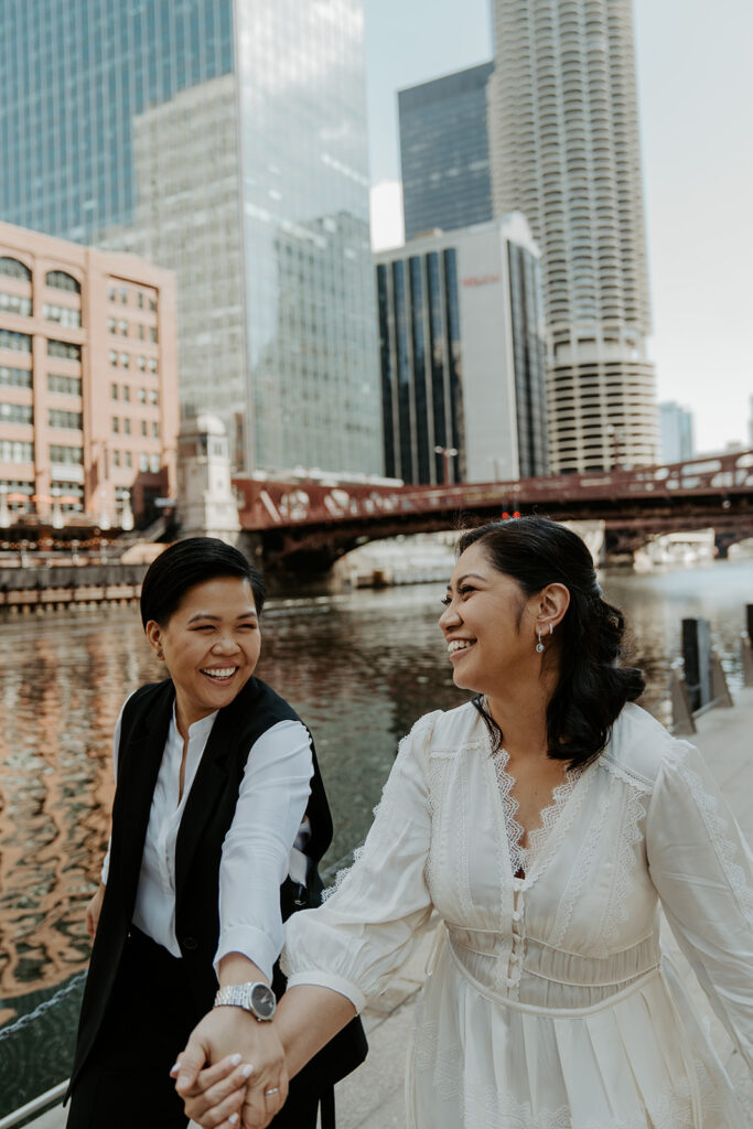 Two brides walk together on the River Walk in Chicago holding hands and smiling at one another.