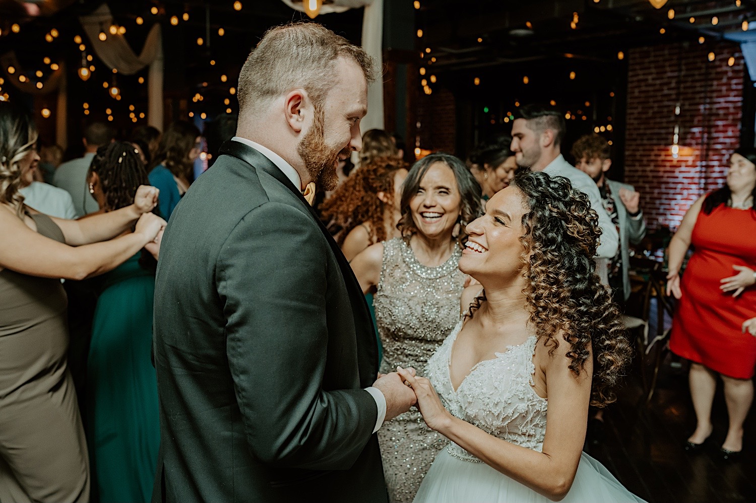 A bride and groom smile at one another while holding hands as they dance with their guests during their wedding reception