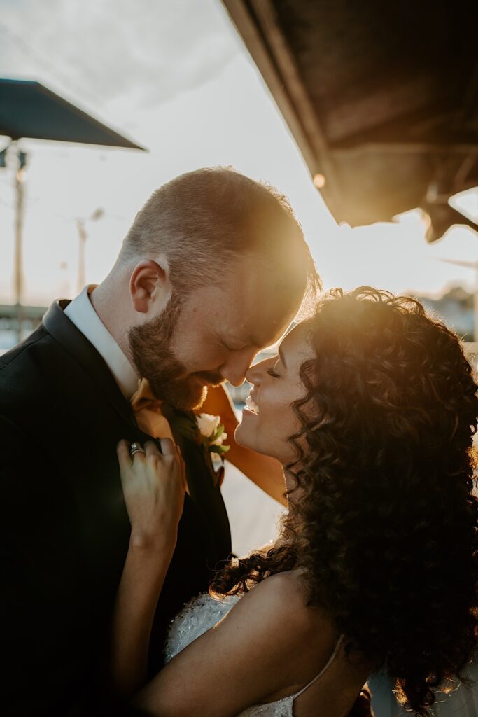 A bride and groom smile as they lean in for a kiss while the sun sets behind them on their wedding day