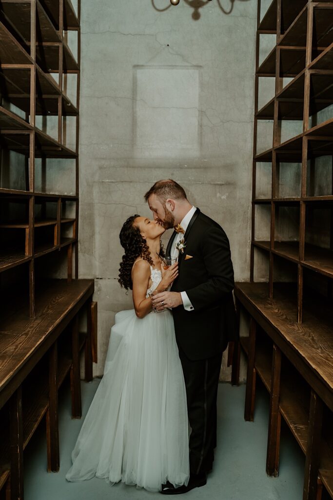 A bride and groom kiss while in a room surrounded by empty wooden shelves as they have their wedding portraits taken