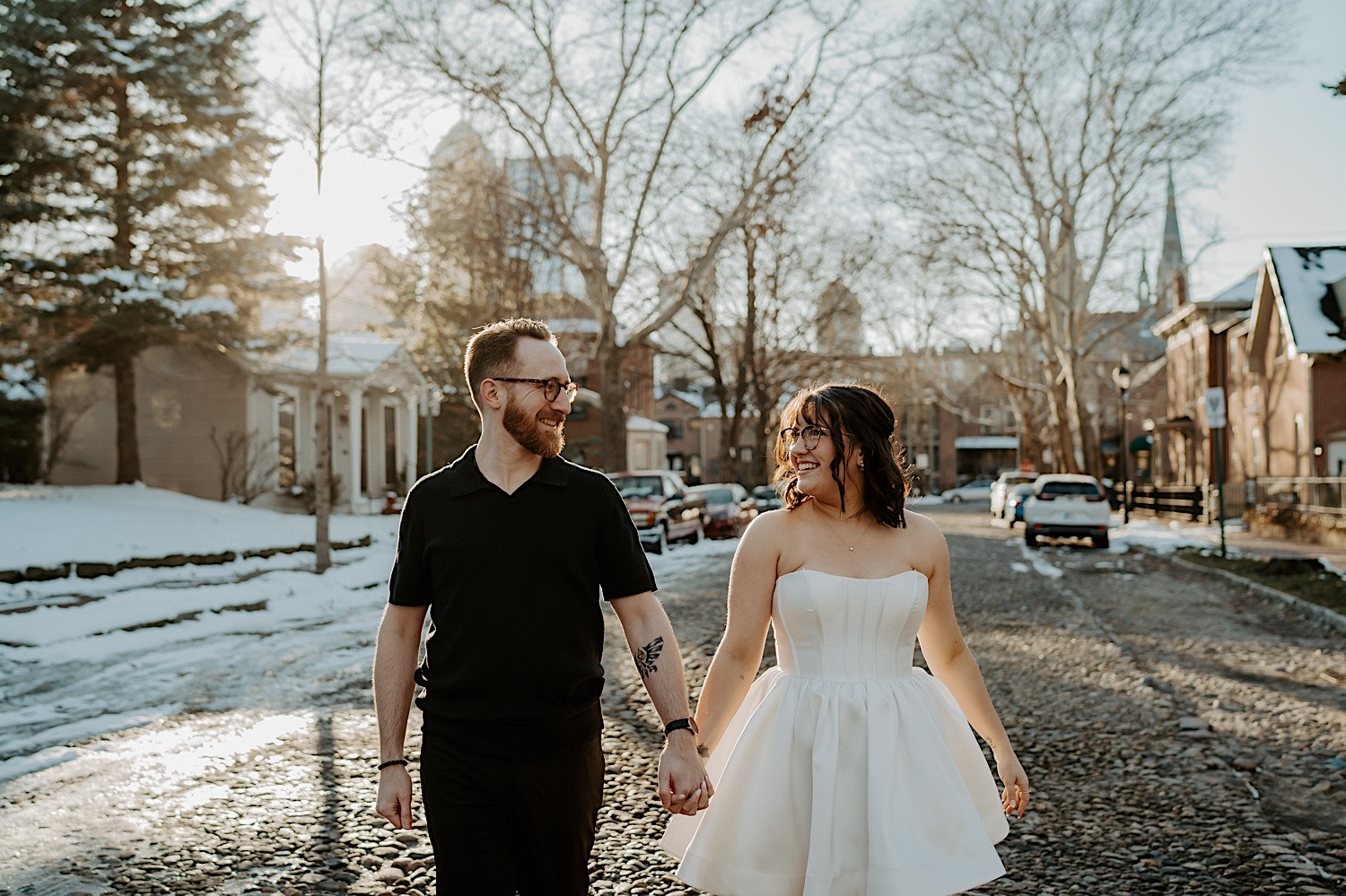 A man and woman walk hand in hand down a snowy street in Indianapolis as they smile at one another while having their anniversary photos taken