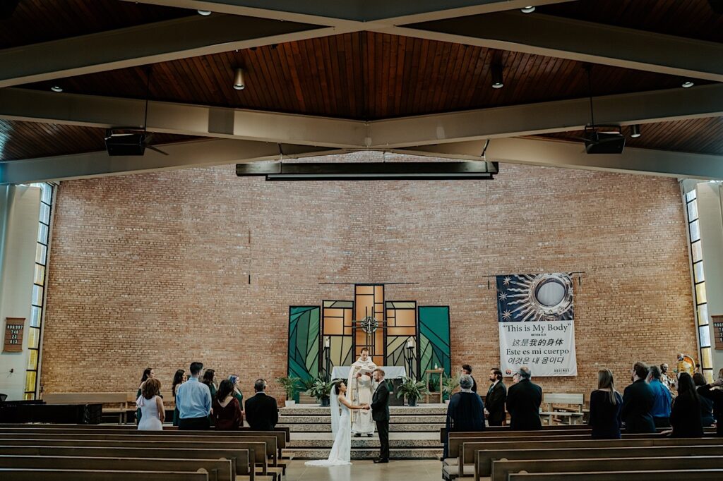A bride and groom stand together in a church as guests stand around them and the pastor speaks during their wedding ceremony