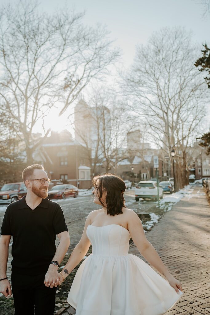 A couple hold hands and smile at one another while on a sidewalk at sunrise