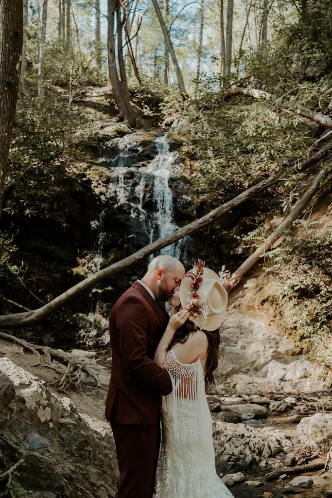 A couple kiss one another while standing in the woods in front of a waterfall as they have their elopement portraits taken