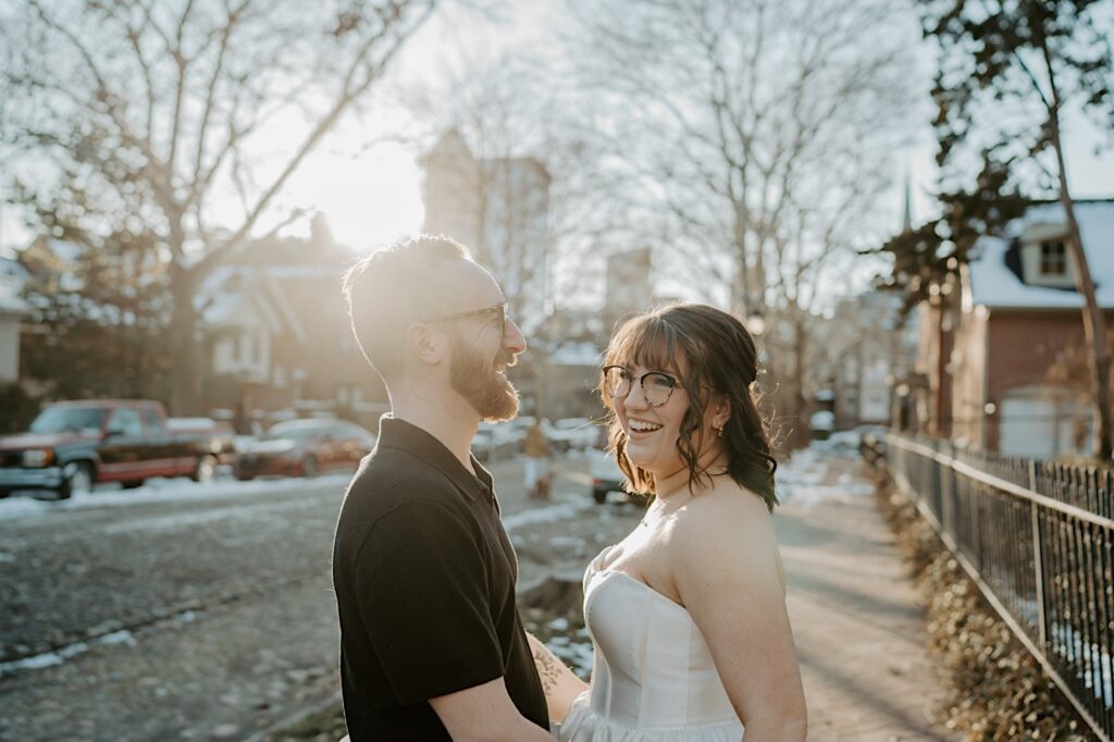 A couple smile while facing one another  as they stand together on a sidewalk in Indianapolis at sunrise while having their anniversary photos taken