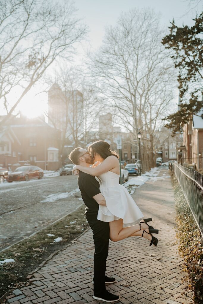 A couple kiss as the man lifts the woman in the air while standing on a sidewalk at sunrise
