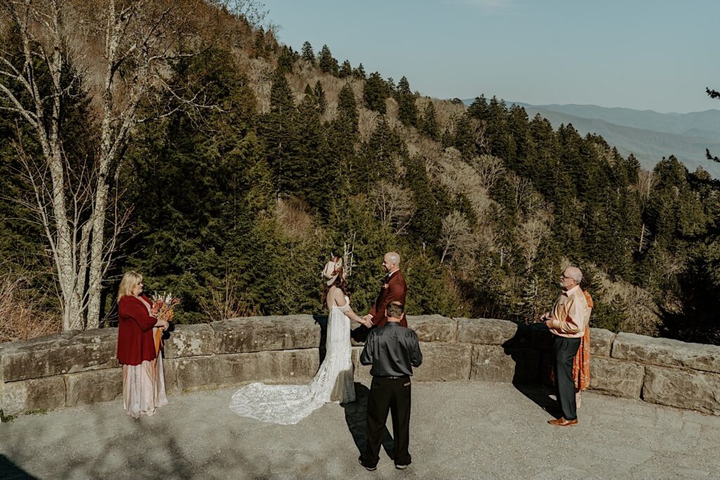 A bride and groom hold hands while their parents stand around them as they have their elopement ceremony on a lookout in the mountains of Tennessee