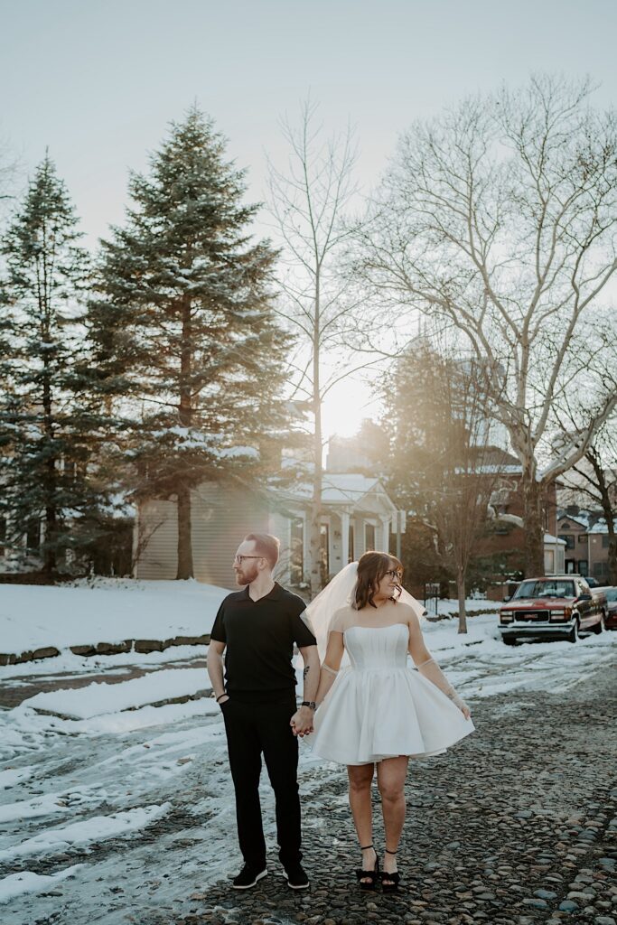 A couple hold hands and look in opposite directions while standing in the middle of a snowy street at sunrise