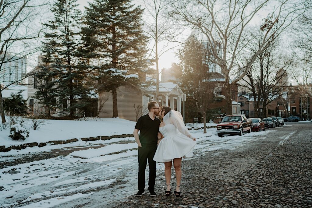 A couple kiss one another while standing in the middle of a snowy street in their neighborhood of Indianapolis as they have their anniversary photos taken