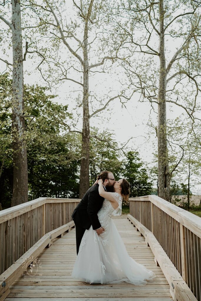 A bride and groom kiss one another while on a wooden bridge in the woods