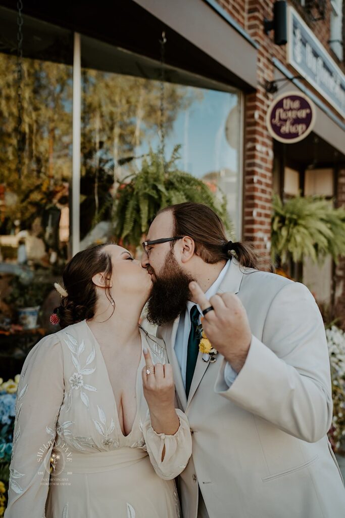 A bride and groom kiss one another  while outside a flower shop as they show off their wedding rings to the camera