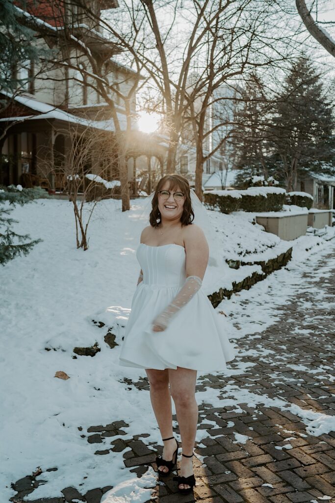 A woman in a white dress smiles at the camera as she stands on a snowy sidewalk at sunrise