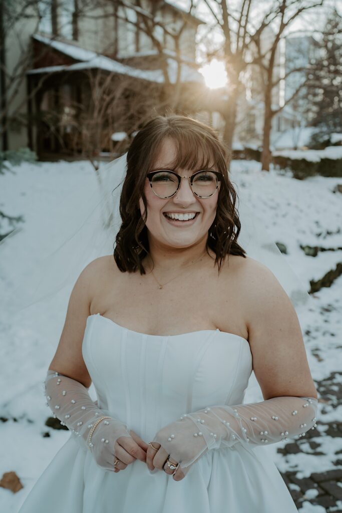 A woman in a white dress smiles at the camera while the sun rises behind her over a snow covered house