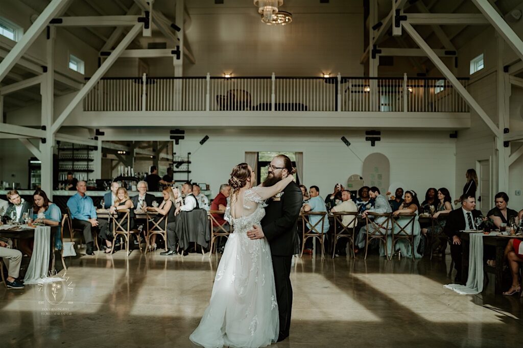 A bride and groom smile at one another while having their first dance together as guests sit and watch during their indoor wedding reception