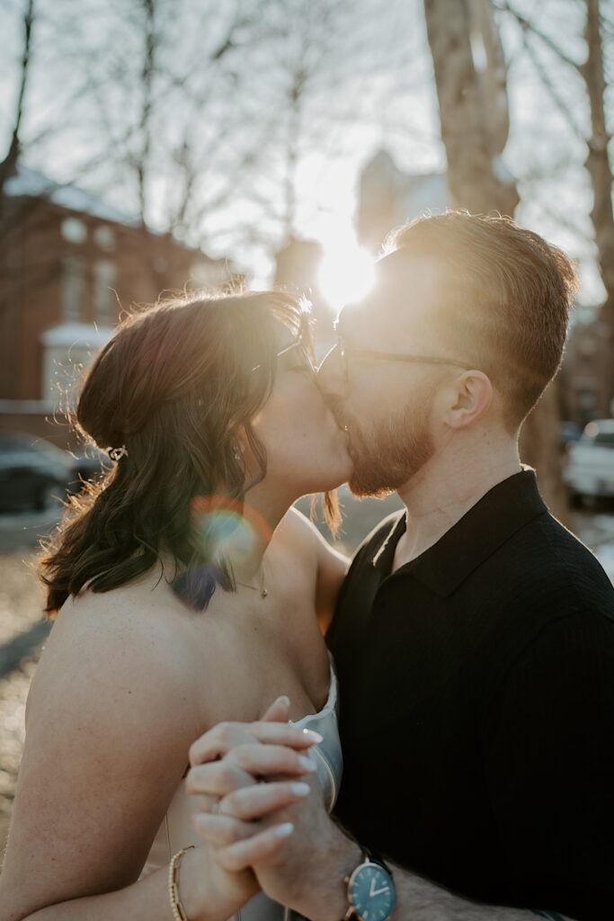 A couple kiss one another while holding hands as the sun rises behind them
