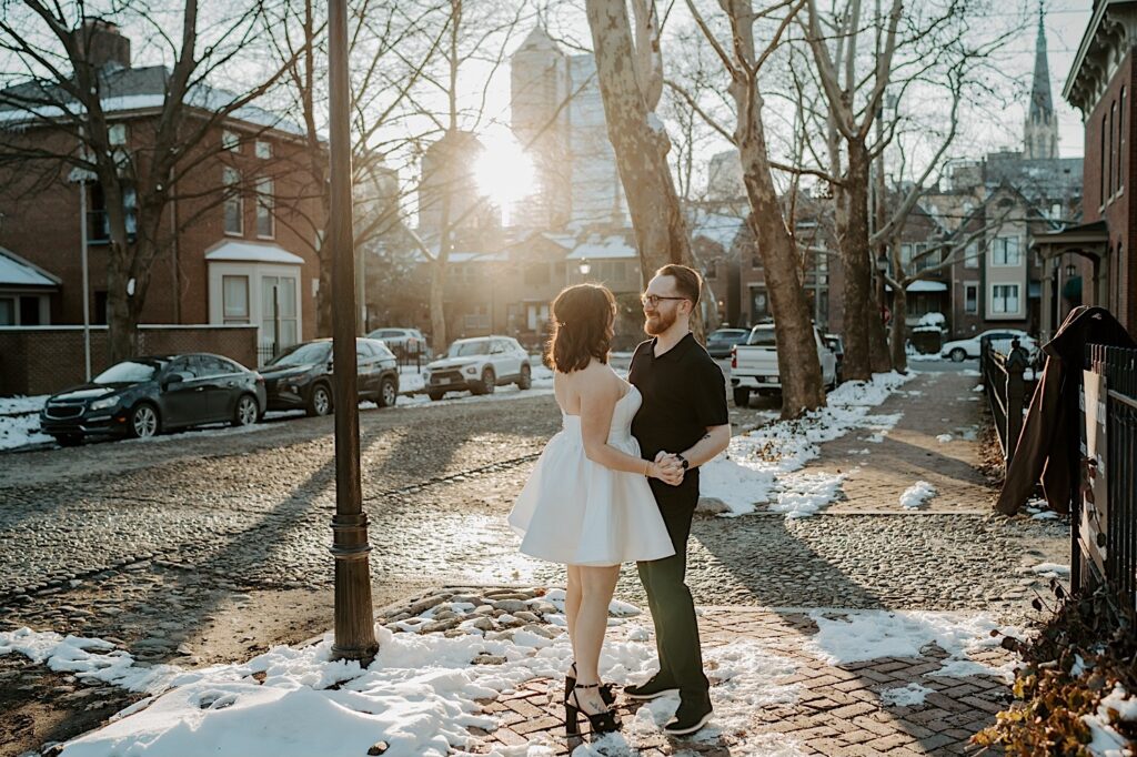 A couple hold hands and face one another while standing on a snow covered sidewalk in Indianapolis as the sun rises while they have their anniversary photos taken