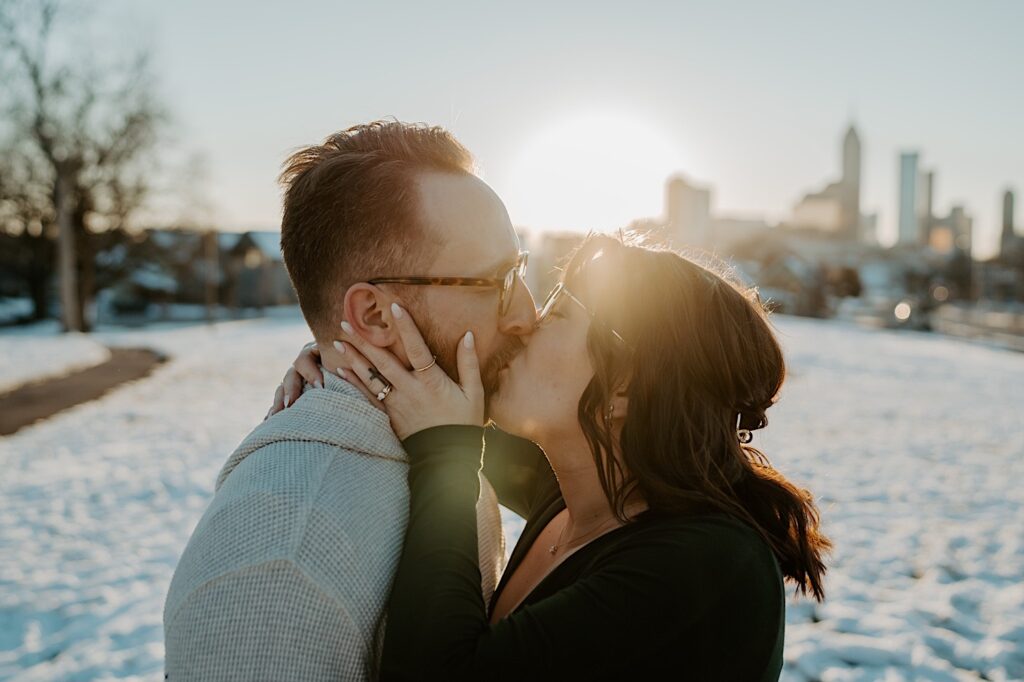 A couple kiss one another while having their anniversary photos taken in a snowy field with the sunrise and the city of Indianapolis behind them 
