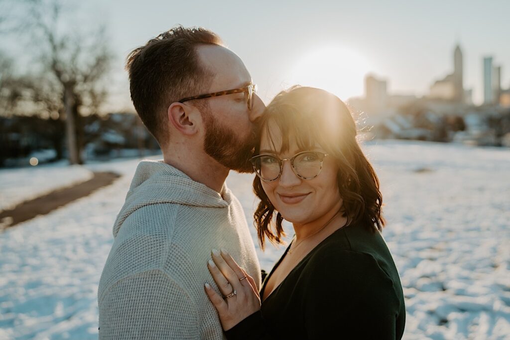 A woman smiles at the camera as a man kisses her head while the two have their anniversary photos taken in a snowy field of Indianapolis at sunrise
