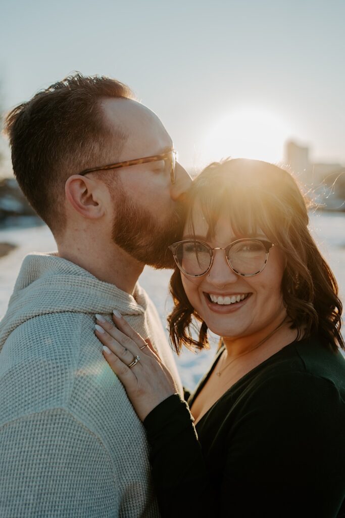 A woman smiles at the camera while a man kisses her head as the sun rises behind them