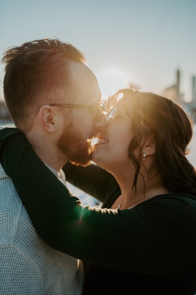 A couple smile as they embrace for a kiss while the sun rises behind them