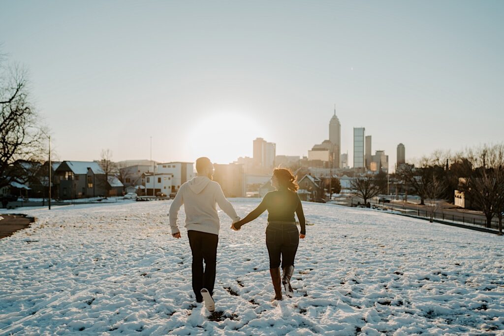A couple hold hands as they run away from the camera through a snowy field towards Indianapolis while having their anniversary photos taken