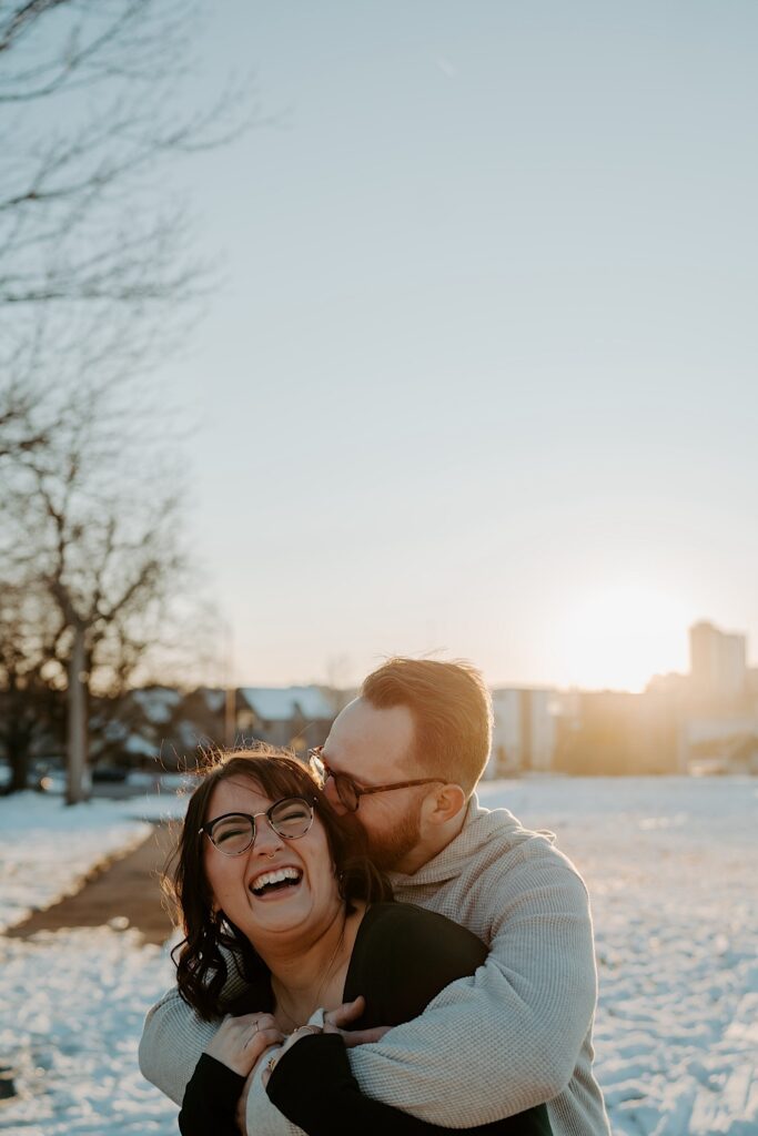 A woman smiles at the camera while a man hugs her from behind and kisses her head while the two stand in a snowy field at sunrise