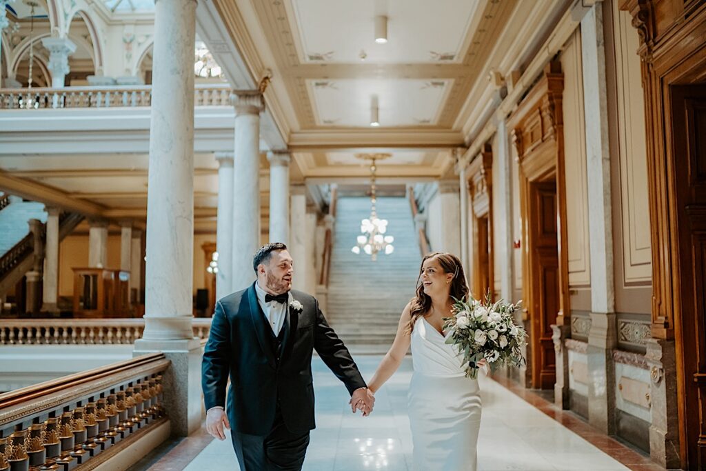 Bride in a white fit dress and a big white floral bouquet holds hands with groom in a black tux and black bowtie in the hallway of the Indiana Statehouse.