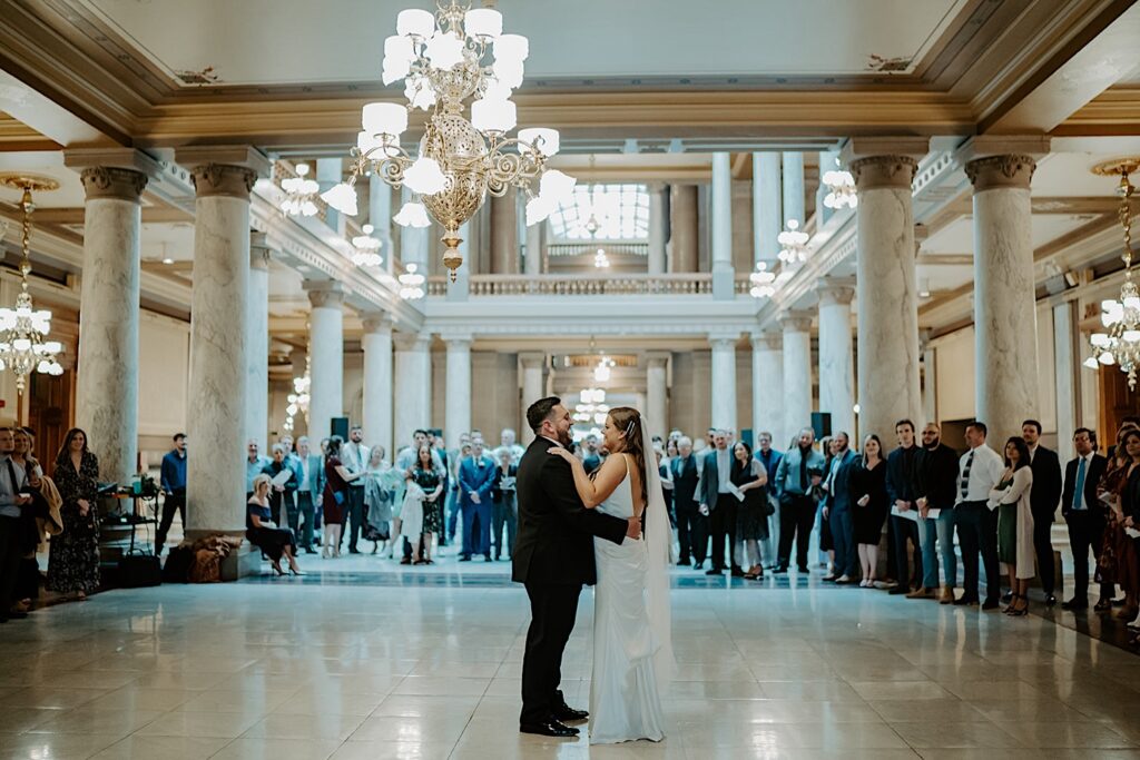 Bride and groom dance in center floor underneath beautiful antique chandeliers surrounded by friends and family at the Indiana Statehouse during wedding reception. 