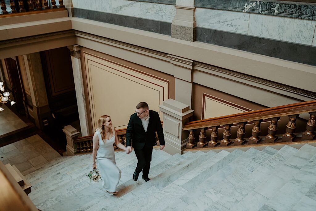 Bride in beautiful white dress holds hands with groom in black suit walking up the stairs at the Indiana Statehouse before ceremony. 