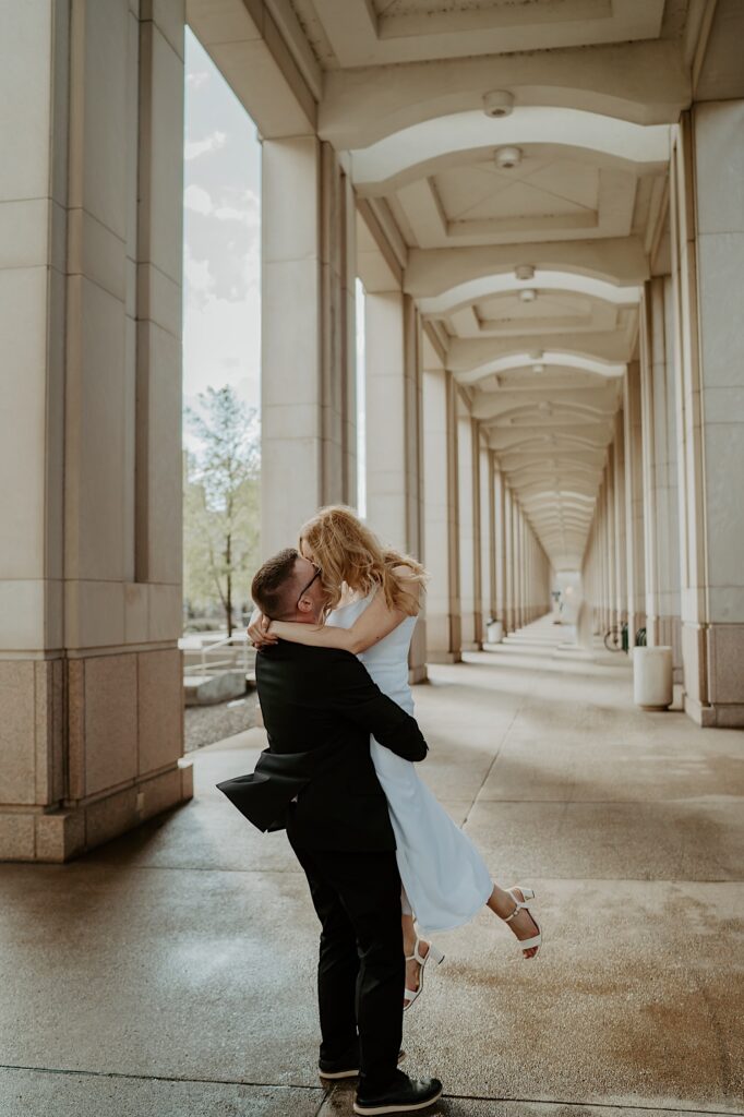Groom in black suit lifts up bride in white dress for a kiss in an outdoor hallway at the Indiana Statehouse. 