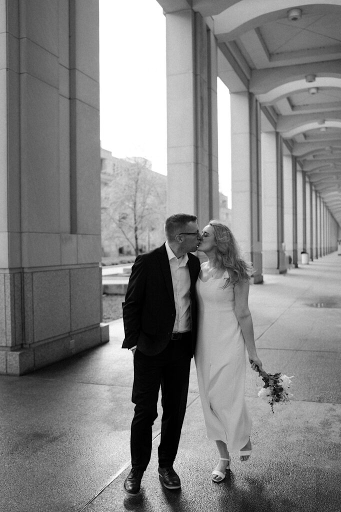 Black and White photo of Groom in black suit kissing Bride in white dress walking down an outdoor hallway at Indiana Statehouse. 