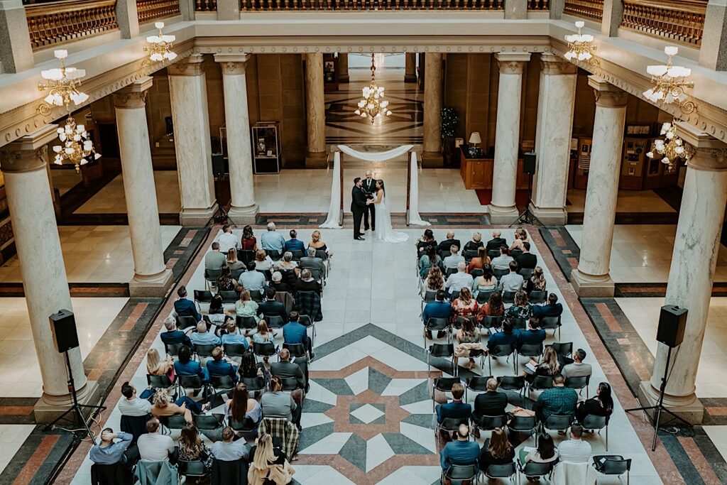 Bird Eye's view of wedding ceremony in the center of Indiana Statehouse with guests sitting on chairs on beautiful marble floor. 