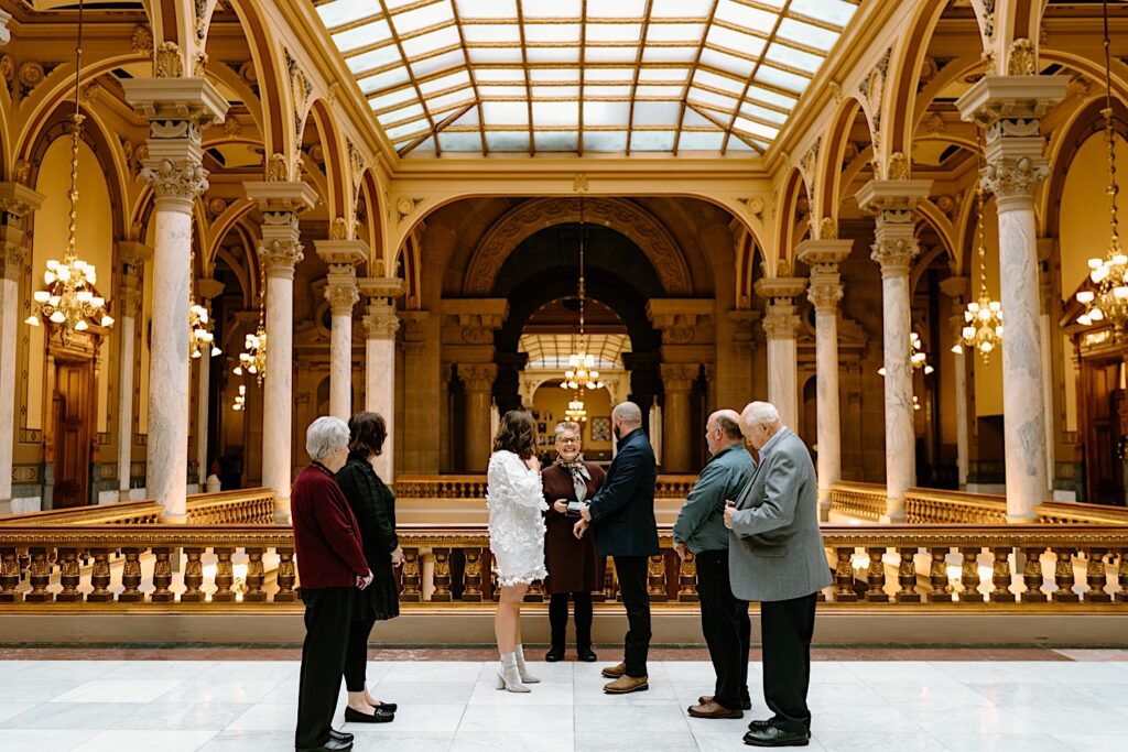 Bride in a short feathery white dress and Groom in a black tux get married on top floor of Indiana Statehouse in an intimate ceremony surrounded by parents of the Bride and Groom. 