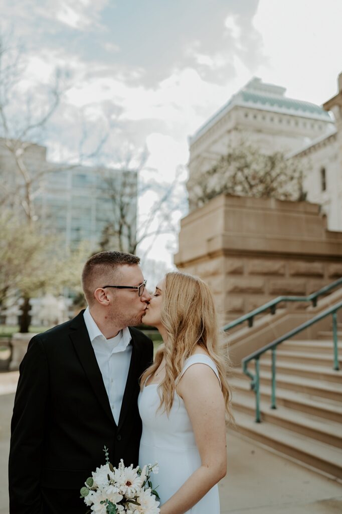 Groom in a black tux and glasses kisses his blonde bride in a white dress with a peachy bouquet outside the steps of the Indiana Statehouse. 
