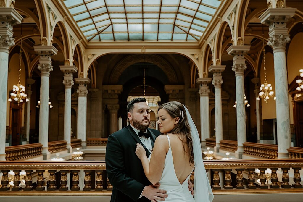 Bride faces her groom showing off her white sheer veil for wedding portraits on the top floor of the Indiana Statehouse surrounded by beautiful gold and marble columns. 