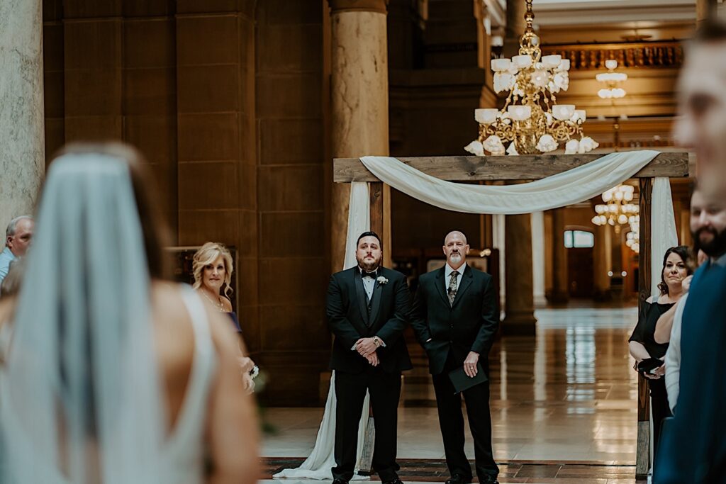 Bride walks down to the end of the aisle to her emotional groom standing underneath an arch and antique gold chandelier in the Indiana Statehouse. 