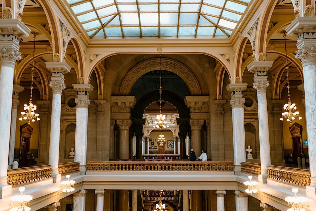 Bride and Groom walk through the top floor of the Indiana Statehouse surrounded by gold and marble columns and beautiful glass ceiling. 