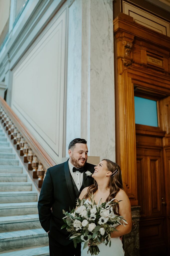 Groom in a black tux and black bow tie looks lovingly at his Bride holding a large white bouquet at the bottom of the steps at the Indiana Statehouse. 