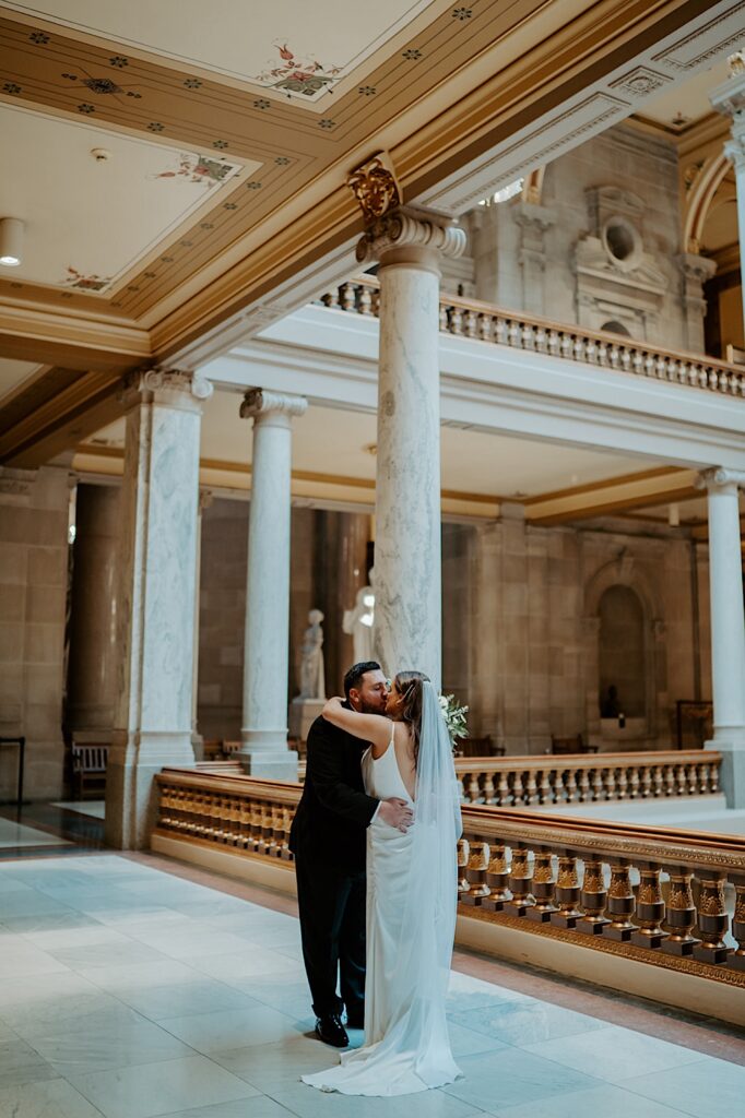Bride and groom kiss near the balcony surrounded by marble columns at the Indiana Statehouse. 