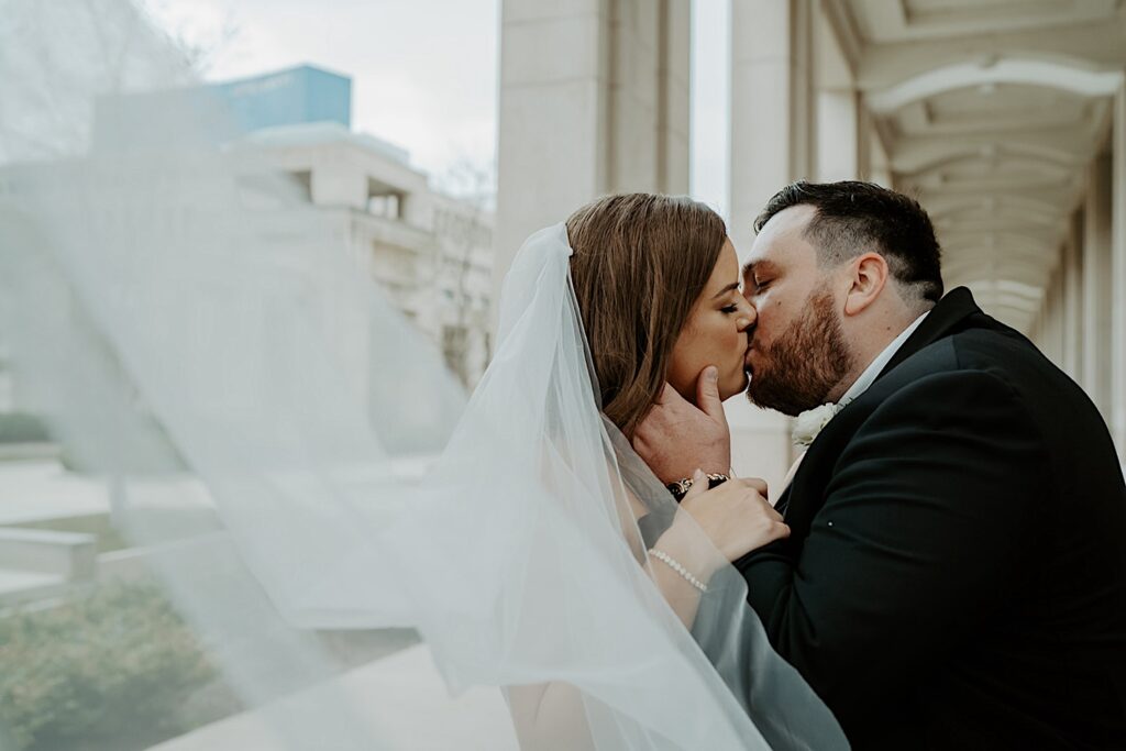 Bride with her veil flowing towards the camera kisses her groom outside underneath an awning at the Indiana Statehouse. 