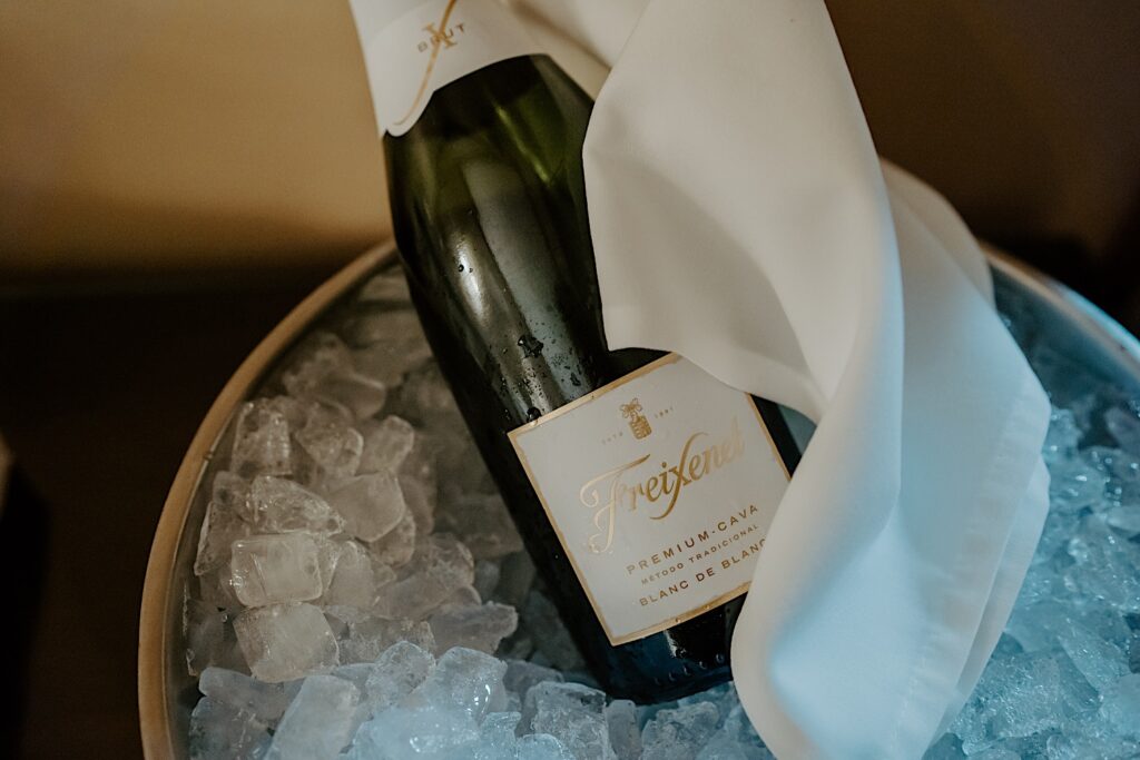 A bottle of Premium Cava Blanc De Blanc sits in a bowl of ice with a napkin wrapped around it. 