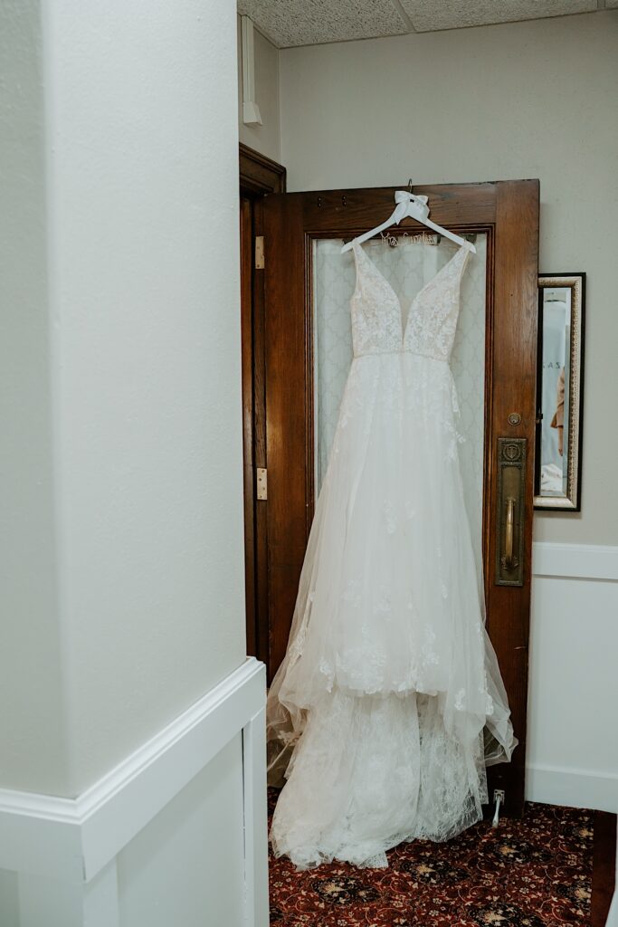 A lacy diamond white low cut wedding dress hangs on a wooden door. 