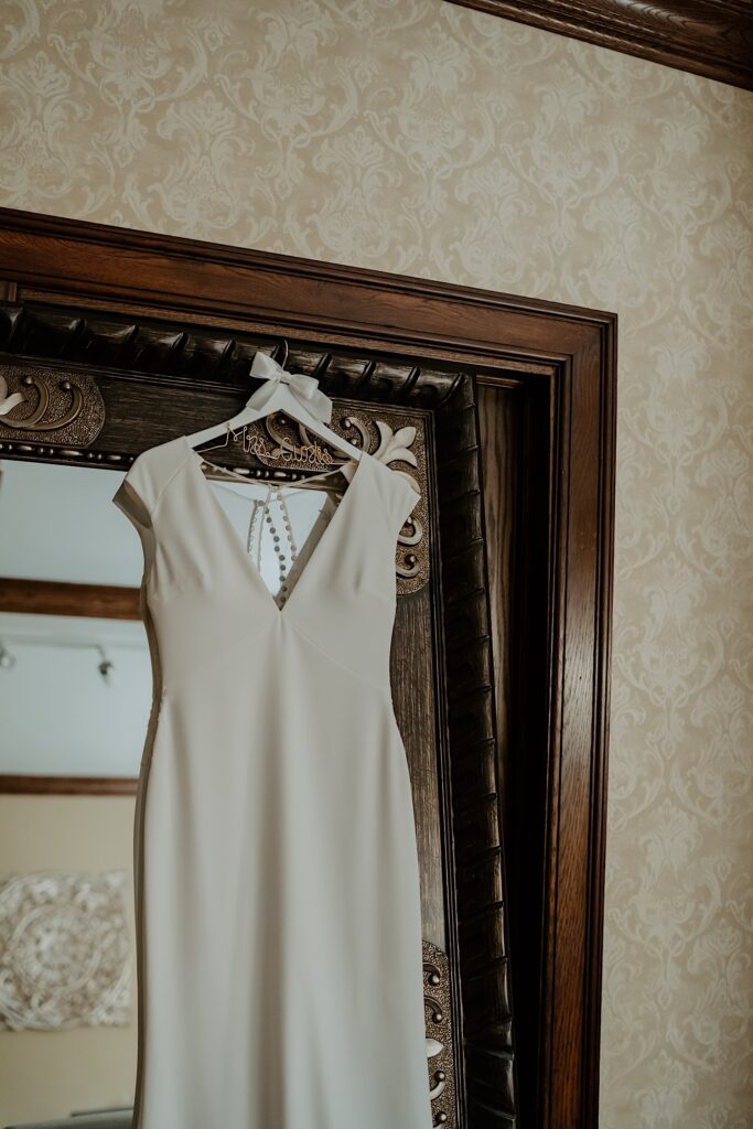 A simple off white gown on a white hanger hangs on a doorway for display.