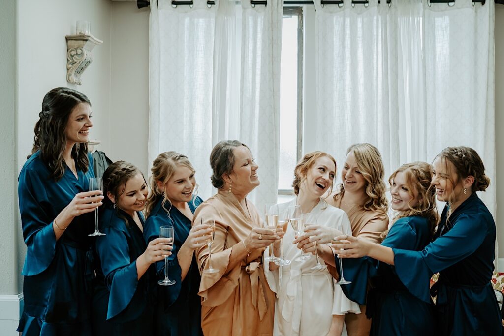 Bride smiles at her bridesmaids candidly in navy blue robes while they hold glasses of champagne towards midwest wedding photographer. 