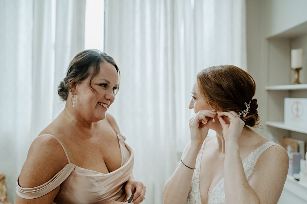 Maid of honor in a champagne bridesmaids dress smiles at bride as she puts pearl earrings in. 