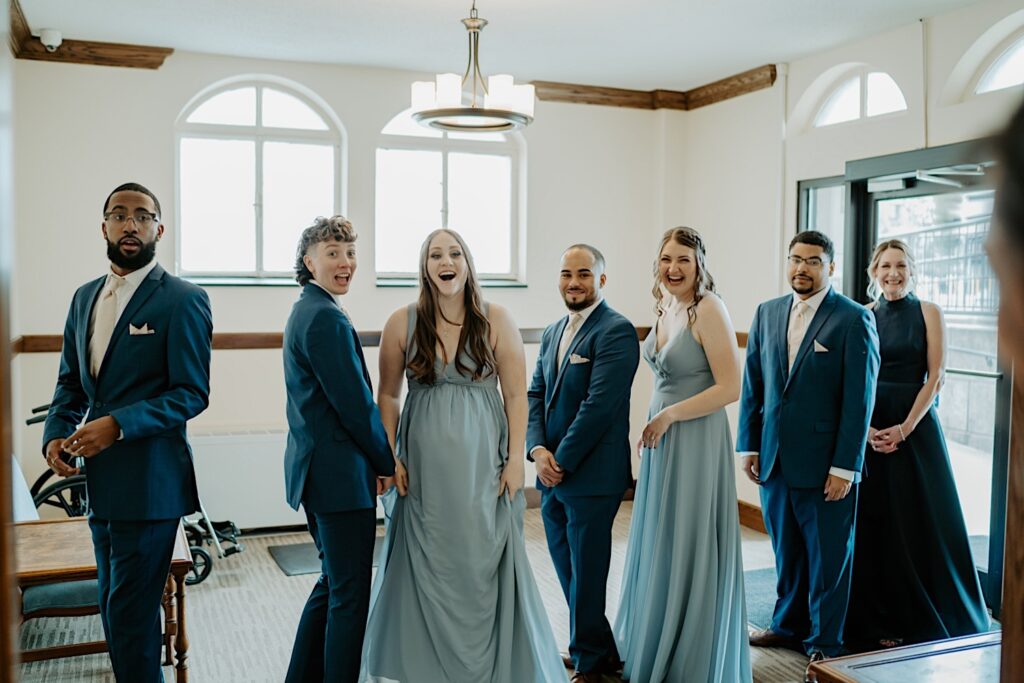 Bridesmaids in dusty blue dresses and groomsmen in blue suits turn around and smile as one of the brides walks out for a first look. 