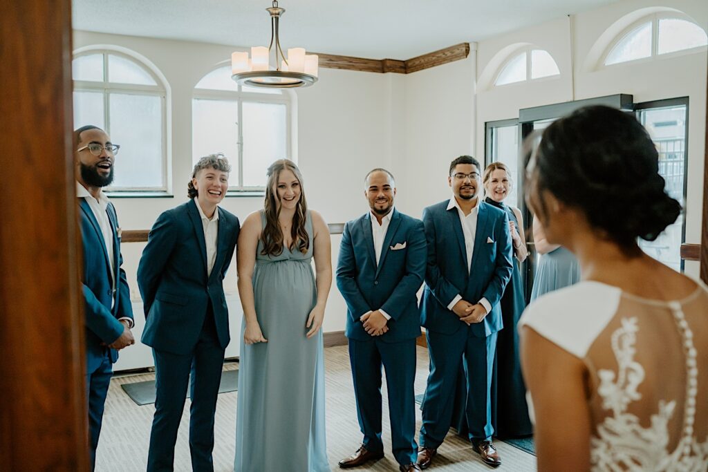 Bridesmaids in dusty blue dresses and groomsmen in blue suits turn around and smile as one of the brides walks out for a first look. 